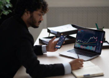 Smart focused Indian broker investor, stock trader using smartphone and laptop to analyze and invert in the financial market of cryptocurrencies, buy or sell crypto coins, planning strategy