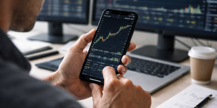 A trader analyzes a financial price chart on a smartphone while multiple market charts display on monitors in the background.