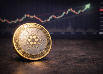 A Cardano (ADA) cryptocurrency token placed on a table with a blurred upward-trending market chart in the background.